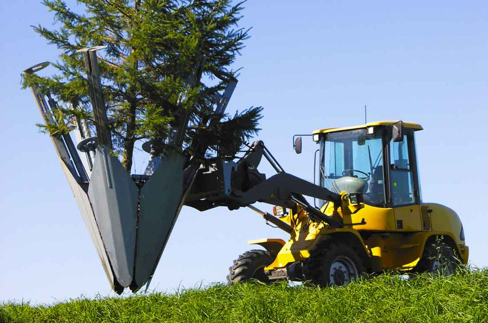A skid steer loader equipped with a Baumalight tree spade attachment, featuring dual hinged blades, is positioned on grass with a clear blue sky in the background.